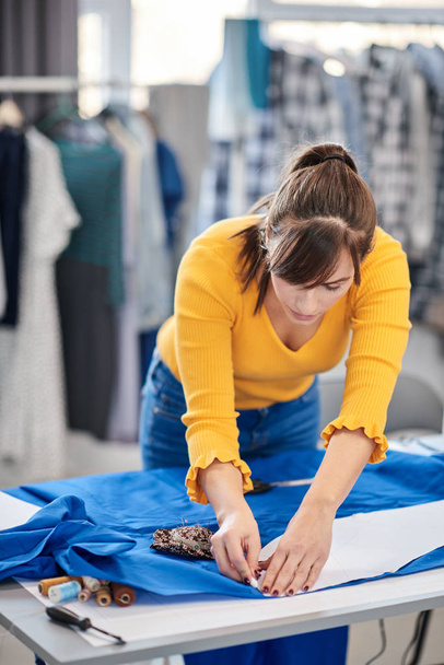 Δημιουργική καυκάσια σχεδιάστρια μόδας στέκεται στο στούντιό της και σχέδιο σε μπλε λινό για ένα όμορφο βραδινό φόρεμα. - Φωτογραφία, εικόνα