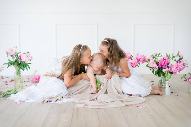 Cute little sisters with blond hair in white dresses and a little brother in a light spring studio decorated with pink and white peonies - Fotoğraf, Görsel