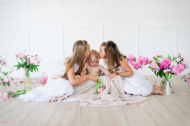 Cute little sisters with blond hair in white dresses and a little brother in a light spring studio decorated with pink and white peonies - Photo, Image