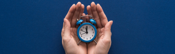 cropped view of woman holding alarm clock on blue background, panoramic shot - Fotoğraf, Görsel