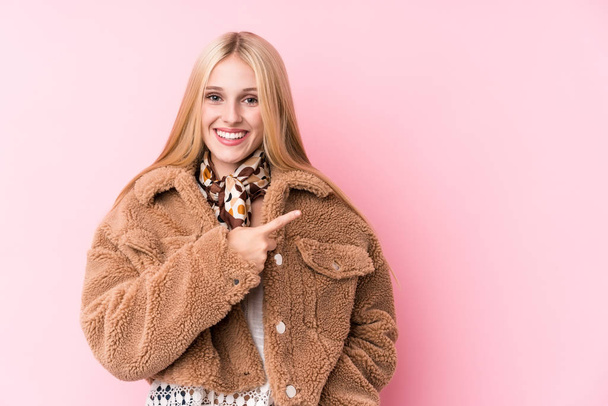 junge blonde Frau im Mantel vor rosafarbenem Hintergrund, lächelnd und zur Seite zeigend, etwas im Leerraum zeigend. - Foto, Bild