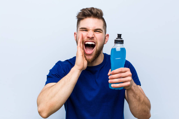Young caucasian sportsman holding an isotonic drink shouting excited to front. - 写真・画像