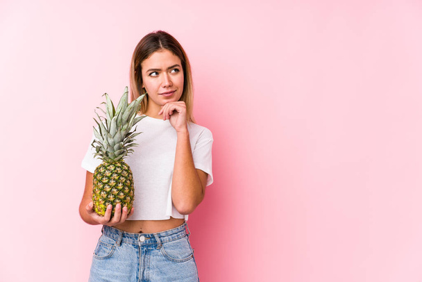 Mulher caucasiana jovem segurando um abacaxi olhando para os lados com expressão duvidosa e cética
. - Foto, Imagem