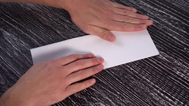 A young man makes an origami plane from a white sheet of office paper on a black textured wooden table. - Imágenes, Vídeo