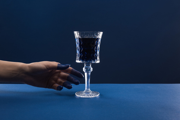 abgeschnittene Ansicht der weiblichen Hand mit bemalten Fingern in der Nähe facettiertes Glas mit Getränk isoliert auf blau - Foto, Bild