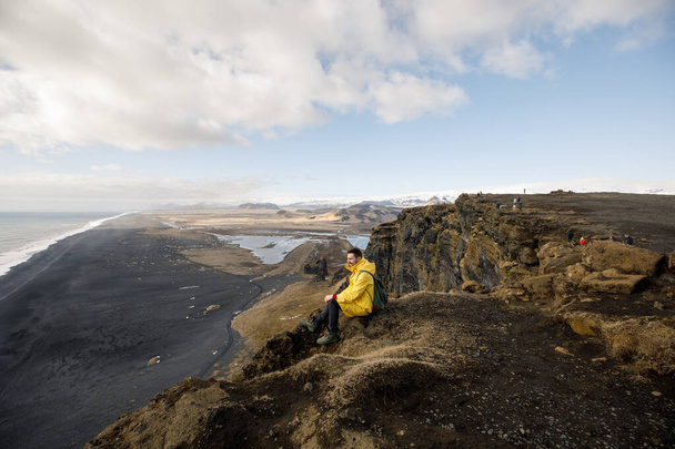 inviting young man on cliff with black sand beach on Iceland mountains view background - Foto, afbeelding