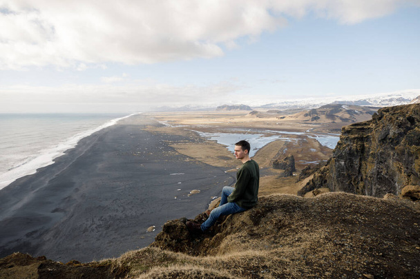 attractive young male sitting on cliff with black sand beach on mountains view background - Foto, afbeelding
