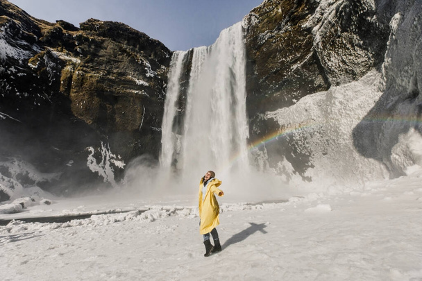 happy woman in yellow coat posing on winter waterfall with rainbow in Iceland mountains background - Zdjęcie, obraz