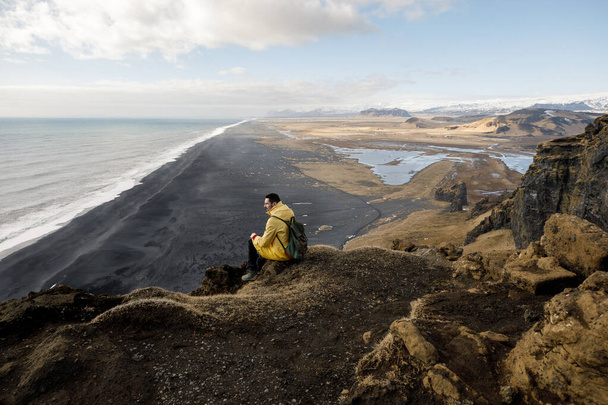 inviting young man on cliff with black sand beach on Iceland mountains view background - Фото, изображение