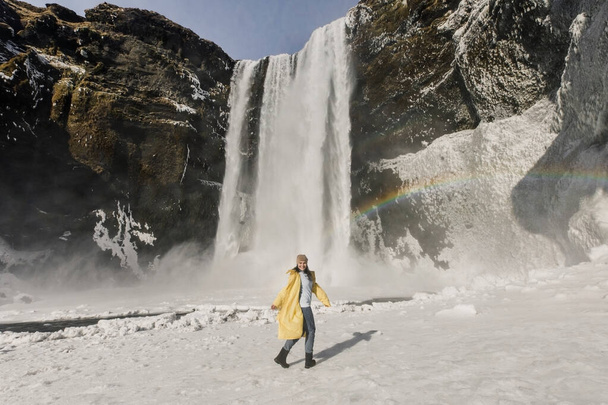 happy woman in yellow coat posing on winter waterfall with rainbow in Iceland mountains background - Fotó, kép