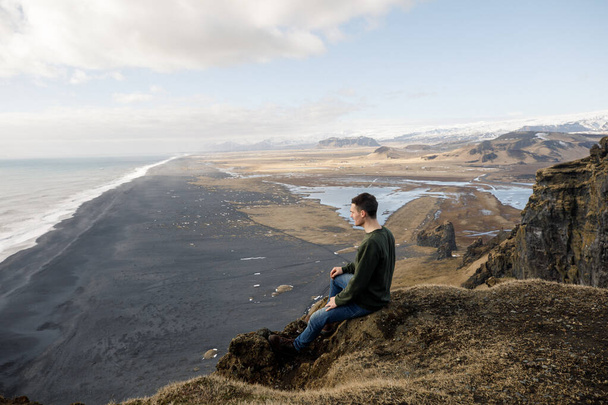 attractive young male sitting on cliff with black sand beach on mountains view background - Photo, Image