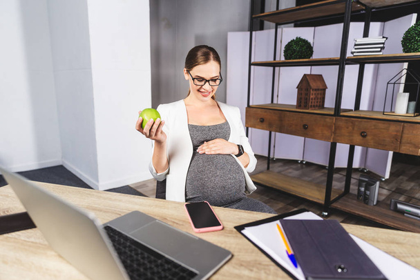 Young pregnant woman holding apple in office - Fotografie, Obrázek