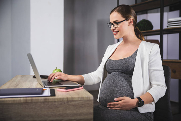 Jovem mulher de negócios grávida usando um laptop no escritório
 - Foto, Imagem