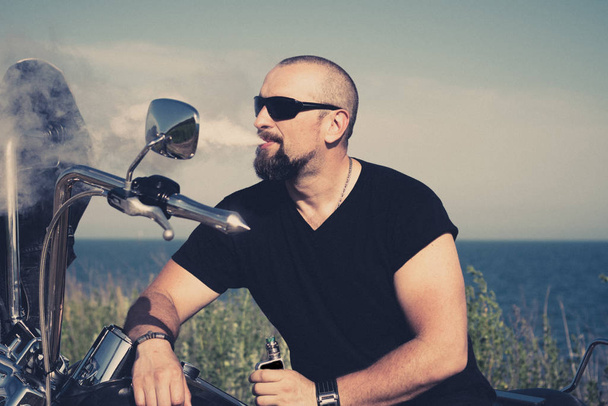 hot biker is smoking sitting on the motorcycle. Brutal man in glasses against the bike - Photo, Image