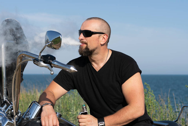 Dögös motoros cigizik a motoron ülve. Brutális férfi szemüvegben a bicikli ellen - Fotó, kép