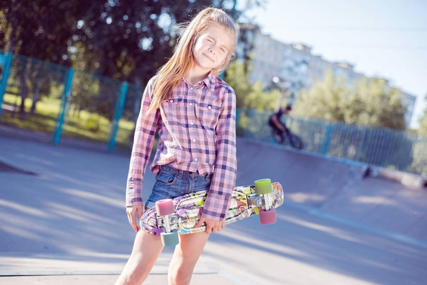 portrait of a beautiful little teenage girl, holding in hand skateboard, in skatepark. on the background rolling around boy on bike - Photo, Image