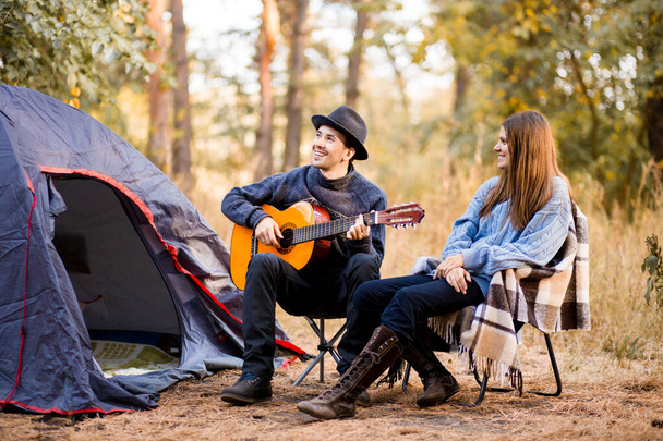Portrait of young couple sitting on camp chair with guitar near camp tent. Time together in harmony with nature far from the bustle of the city - Fotoğraf, Görsel