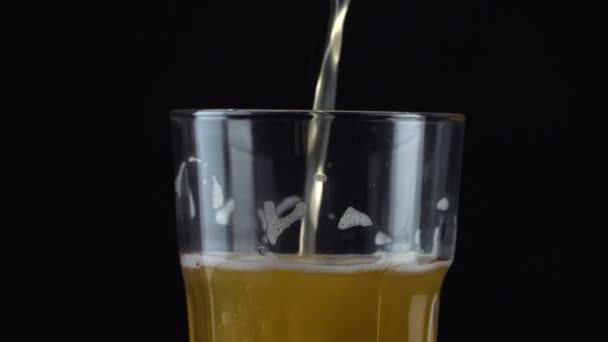 Pouring fresh and cold craft beer into a glass with white foam on top on black background. Flowing foamy wheat or lager beer on dark background - Video, Çekim