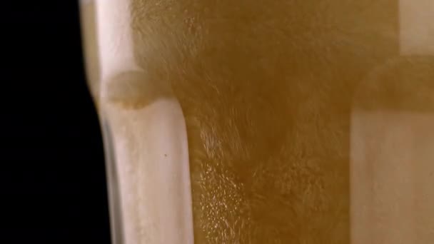 Zblízka čerstvé a studené řemeslné pivo ve sklenici s bílou pěnou na černém pozadí. Makro záběr tekoucí pěny pšenice nebo ležáku piva na tmavém pozadí - Záběry, video
