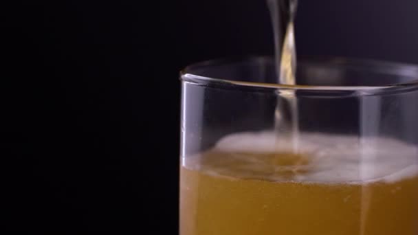 Close up of fresh and cold craft beer in a glass with white foam on top on black background. Macro shot of flowing foamy wheat or lager beer on dark background - Footage, Video
