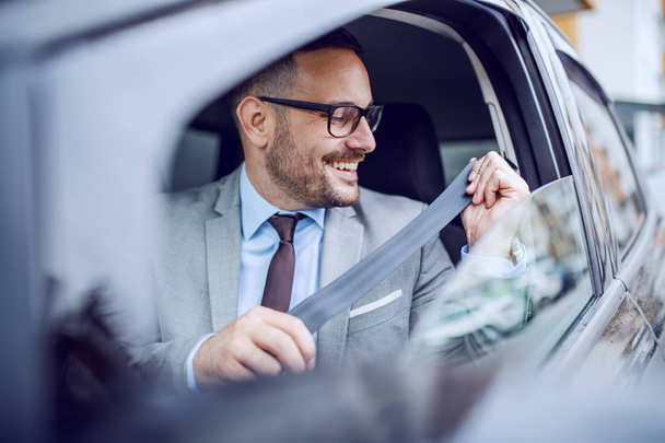 Atraktivní nóbl běloch usmívající se obchodník v obleku a s brýlemi zapínající bezpečnostní pás v autě. - Fotografie, Obrázek