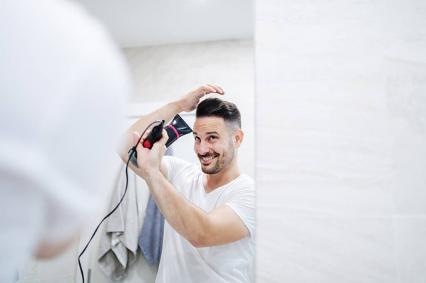 Lustrzane odbicie atrakcyjnego białego mężczyzny w białej koszulce stojącego w łazience i suszącego włosy suszarką do włosów. Poranna rutyna. - Zdjęcie, obraz