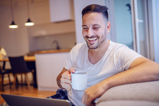 Όμορφος χαμογελαστός αξύριστος καυκάσιος άντρας με πιτζάμες κάθεται στο σαλόνι με φορητό υπολογιστή στην αγκαλιά και πίνει τον φρέσκο πρωινό καφέ του. - Φωτογραφία, εικόνα