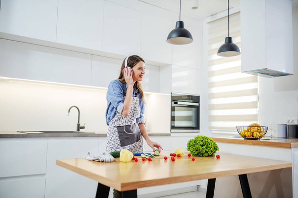 Jovem mulher caucasiana feliz em avental em pé na cozinha e ouvir música. No balcão da cozinha estão todos os tipos de legumes. Conceito de alimentação saudável
. - Foto, Imagem