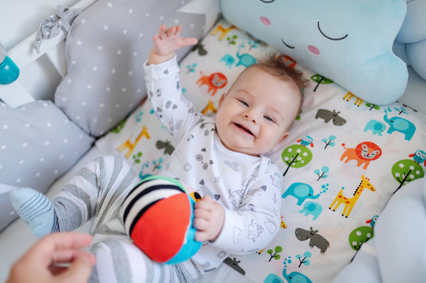 Top view of adorable Caucasian little baby boy with blue eyes lying in crib, catching stuffed ball and looking at camera. - Photo, Image