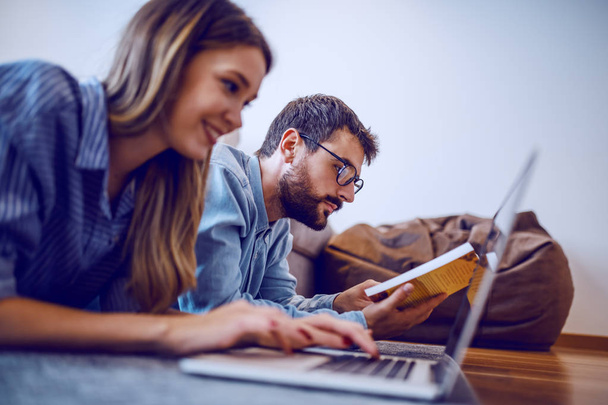 Side view of handsome caucasian bearded man with eyeglasses reading book while his girlfriend smiling, lying next to him and using laptop. Selective focus on man. - Foto, Imagen
