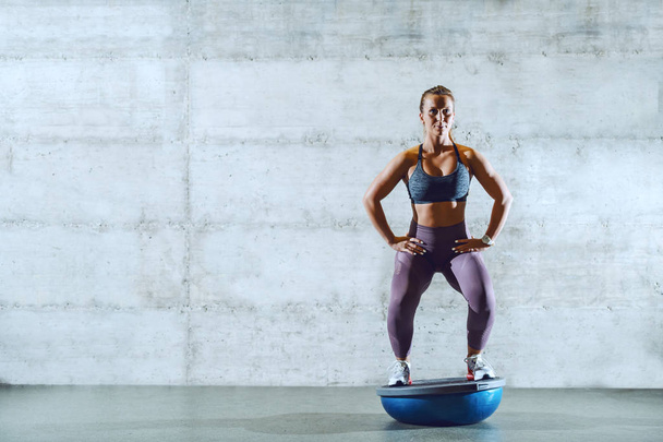 Full length of attractive young caucasian sportswoman in sportswear with ponytail standing on bosu ball and doing squat endurance, - Фото, зображення