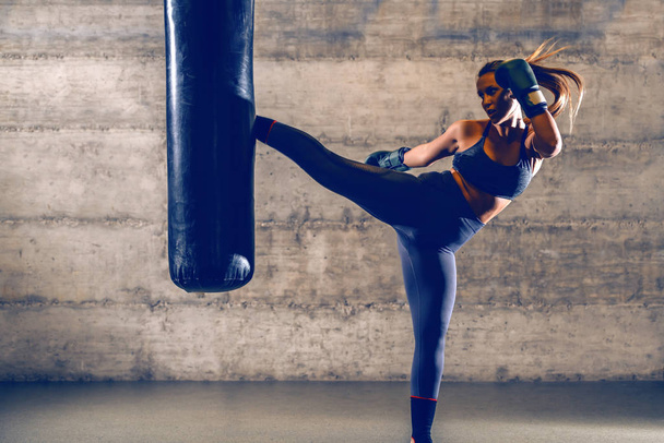 Joven ajuste atractivo musculoso caucásico mujer kick boxer en ropa deportiva con guantes de boxeo patadas bolsa de boxeo
. - Foto, imagen
