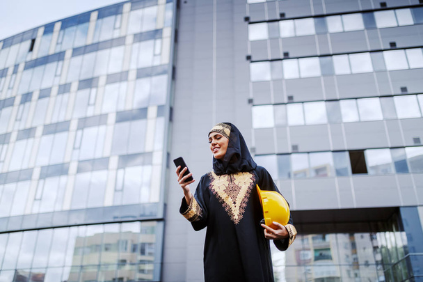Atractiva mujer musulmana sonriente de pie frente al edificio corporativo, usando un teléfono inteligente y sosteniendo el casco bajo la axila. Las mujeres también pueden ser grandes arquitecturas.
. - Foto, Imagen