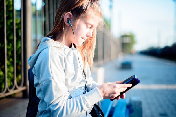 kleines Mädchen, das über Kopfhörer Musik hört und mit dem Smartphone in der Hand auf den Zug wartet - Foto, Bild