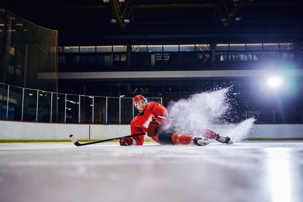 Όμορφος καυκάσιος νεαρός παίκτης χόκεϊ έπεσε στον πάγο κατά τη διάρκεια του αγώνα. - Φωτογραφία, εικόνα