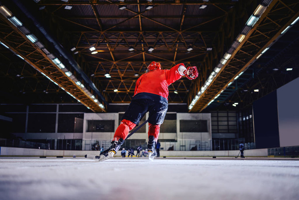 Vista trasera del jugador de hockey patinando hacia el equipo opuesto y tratando de anotar. Hall interior
. - Foto, Imagen