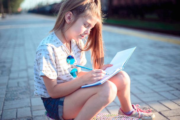 Teenager-Mädchen sitzt auf einem Skateboard und schreibt in ein Notizbuch - Foto, Bild