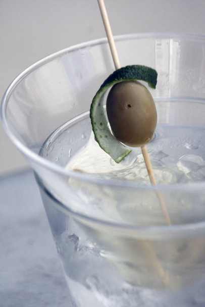 Egy pohár átlátszó alkoholtartalmú ital olívaolajjal, lime héjjal és jégkockával.Üveg közeli kép jégkockákkal, olívával és lime-mal - Fotó, kép
