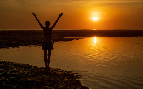 Nadšená mladá žena zvedá paže na pláži před oceánem. Pohled zezadu. Zlatá hodina západu slunce na pláži. Bali, Indonésie. - Fotografie, Obrázek