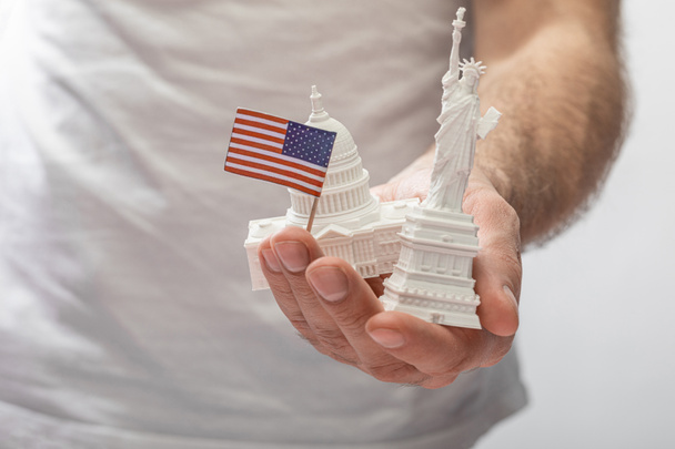 vista recortada del hombre sosteniendo una pequeña estatua de la libertad y el capitolio de los estados unidos cerca de una pequeña bandera americana aislada en blanco
  - Foto, imagen