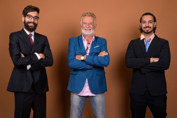 Studioaufnahme von drei bärtigen Geschäftsleuten mit multiethnischem Hintergrund vor braunem Hintergrund - Foto, Bild