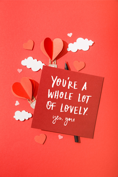 top view of paper heart shaped air balloons in clouds near card with you're a whole lot of lovely yes, you lettering on red background - Photo, Image
