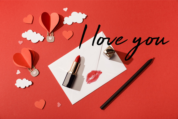 top view of paper clouds and heart shaped air balloons, lipstick and pencil near envelope with lip print and i love you lettering on red background - Photo, Image