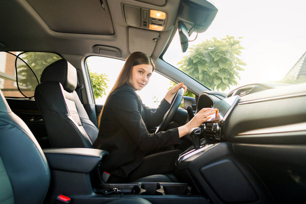 Beautiful smiling young woman in black business suit driving car, attractive girl sitting in automobile, outdoors summer portrait. - Zdjęcie, obraz