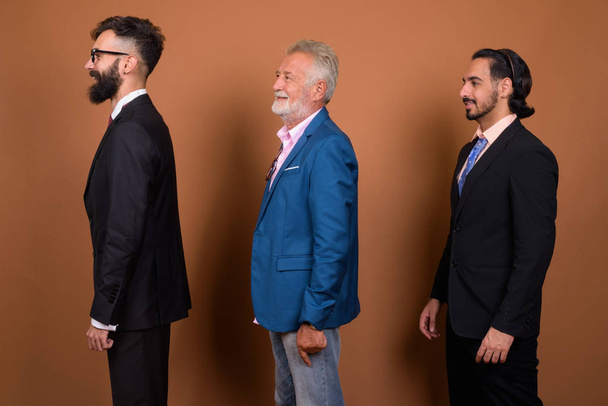 Studioaufnahme von drei bärtigen Geschäftsleuten mit multiethnischem Hintergrund vor braunem Hintergrund - Foto, Bild