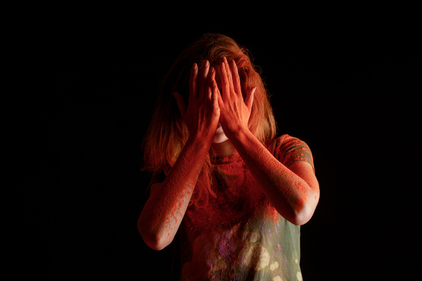 женщина покрывает лицо оранжевой красочной краской цвета голи на руках на черном фоне
 - Фото, изображение