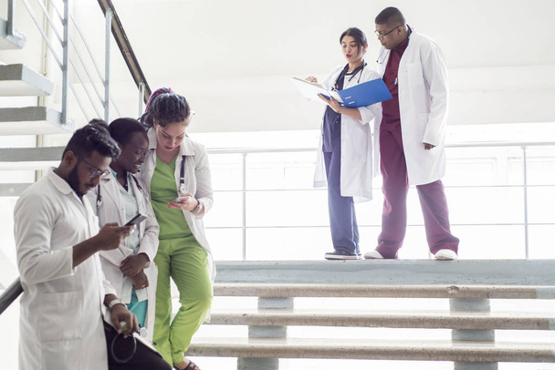 een groep dokters op de trappen in het ziekenhuis. Jongeren van gemengde ras, in medische kleding, onderzoeken röntgenfoto 's, gebruik maken van een smartphone - Foto, afbeelding