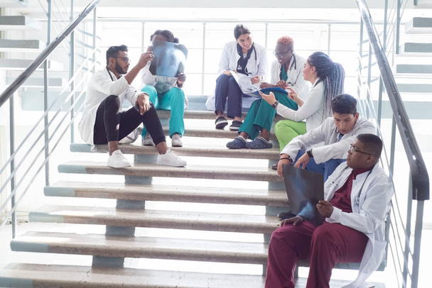 Ärzte sitzen im Krankenhaus auf der Treppe. Junge Menschen unterschiedlichen Geschlechts, gemischte Rassen, in medizinischer Kleidung, mit Phonendoskopen. Krankenakten untersuchen, Smartphone benutzen, Röntgenbilder untersuchen - Foto, Bild