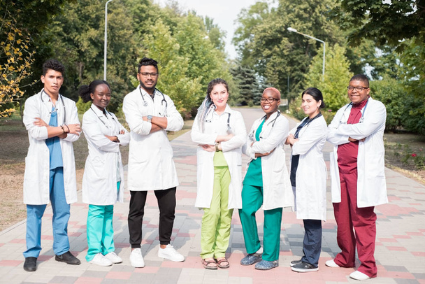 Team gemischter Rennärzte. Gruppe junger Menschen unterschiedlichen Geschlechts in farbiger medizinischer Kleidung, auf der Straße stehend, auf der Straße, mit Phonendoskopen, posierend, lächelnd - Foto, Bild