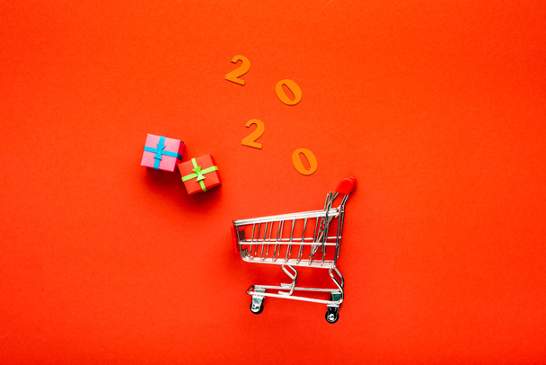 top view of decorative small shopping cart near gift boxes and number 2020 on red background - Photo, Image
